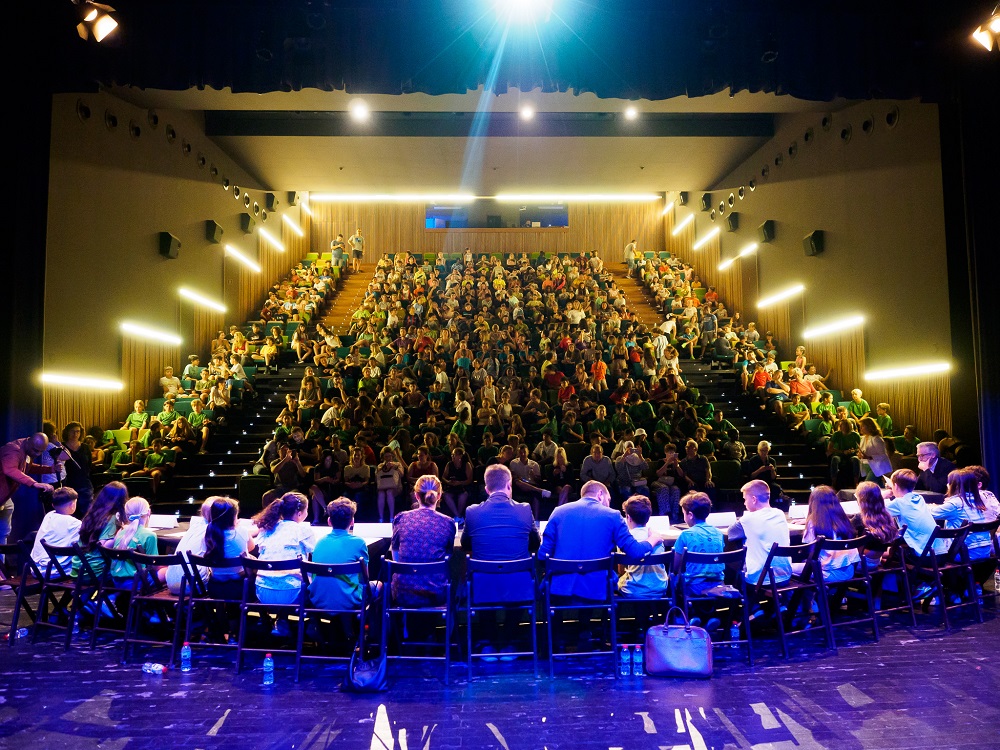 Sala de conferències amb persones adultes i infants a l'escenari que es veuen d'esquenes i l'audiència a davant